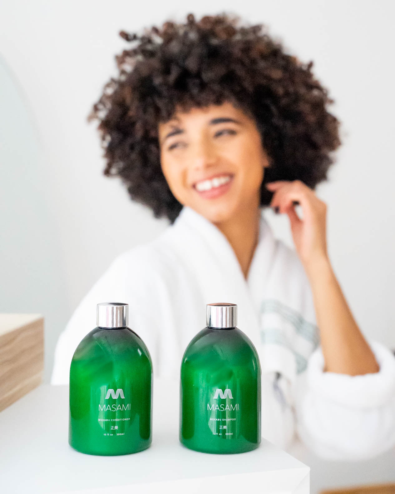 Woman with curly hair wearing a white robe, sitting behind two green bottles labeled 'MASAMI' on a white surface.