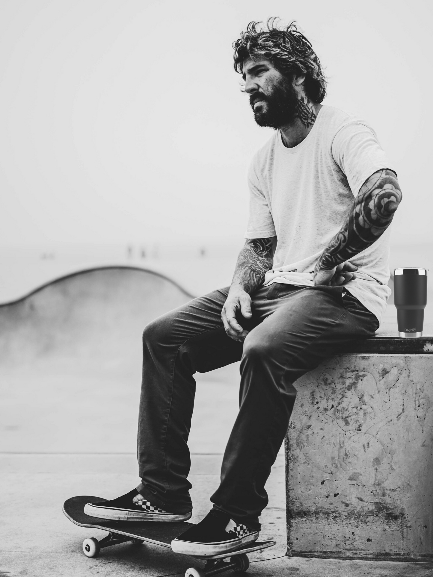 Man with tattoos sitting on a ledge with a skateboard, wearing a white t-shirt and dark pants with his DrinCo Tumbler.