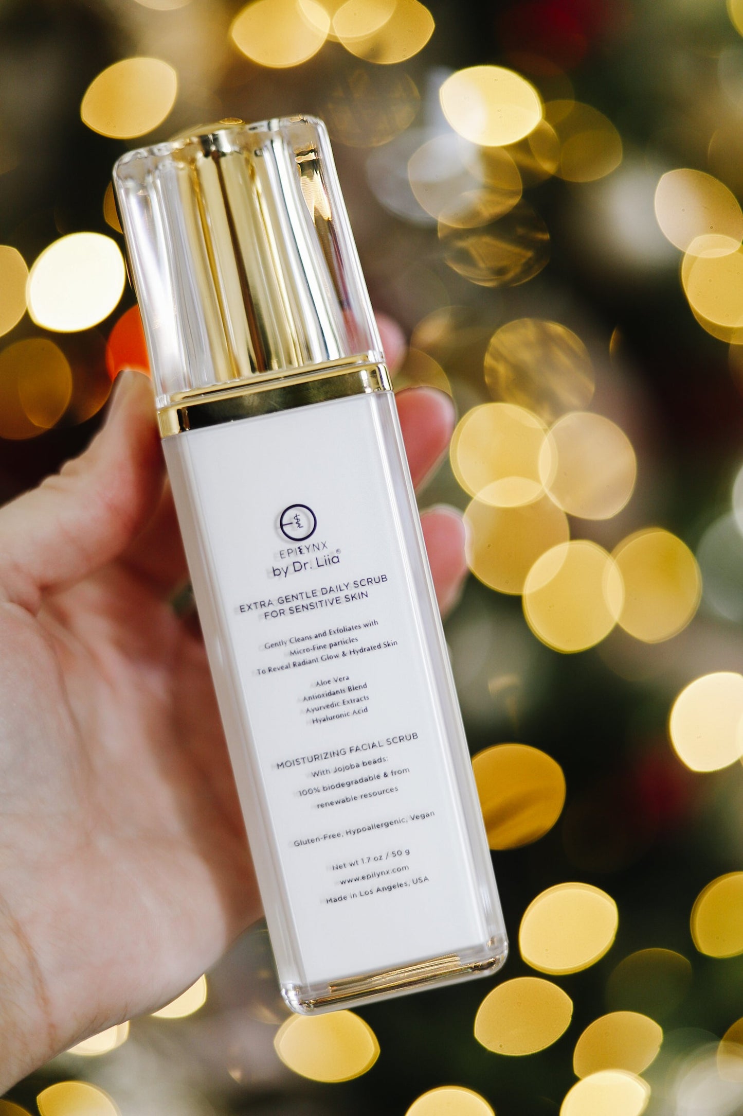 Hand holding EpiLynx by Dr. Liia Extra Gentle Daily Scrub — Hyaluronic Acid, Aloe Vera & Biodegradable Jojoba Beads for Sensitive Skin bottle with a blurred festive background