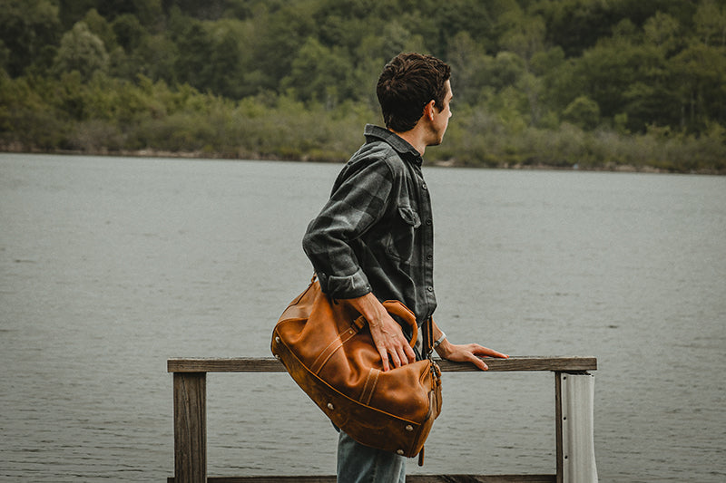Man with a brown leather bag standing on a wooden dock by a lake