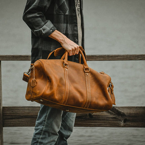 Person holding a brown leather duffel bag against a wooden fence.