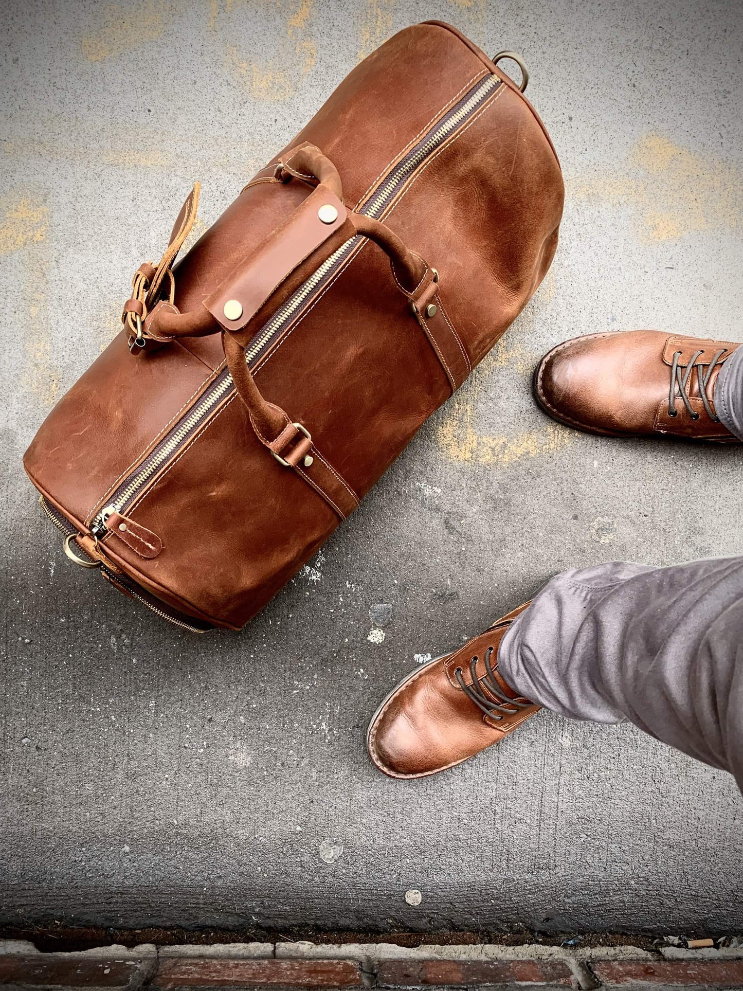Brown leather duffel bag on a textured surface with a person wearing brown shoes.