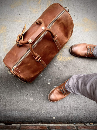 Brown leather duffel bag on a textured surface with a person wearing brown shoes.