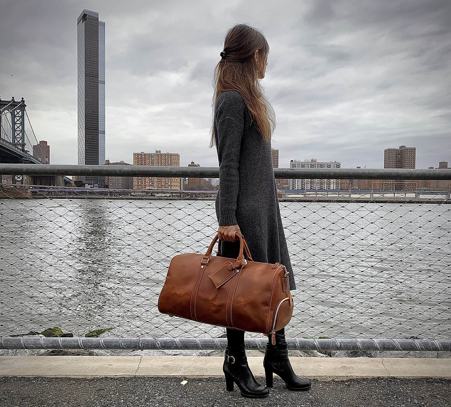 Woman holding a brown leather bag by a waterfront with a city skyline in the background