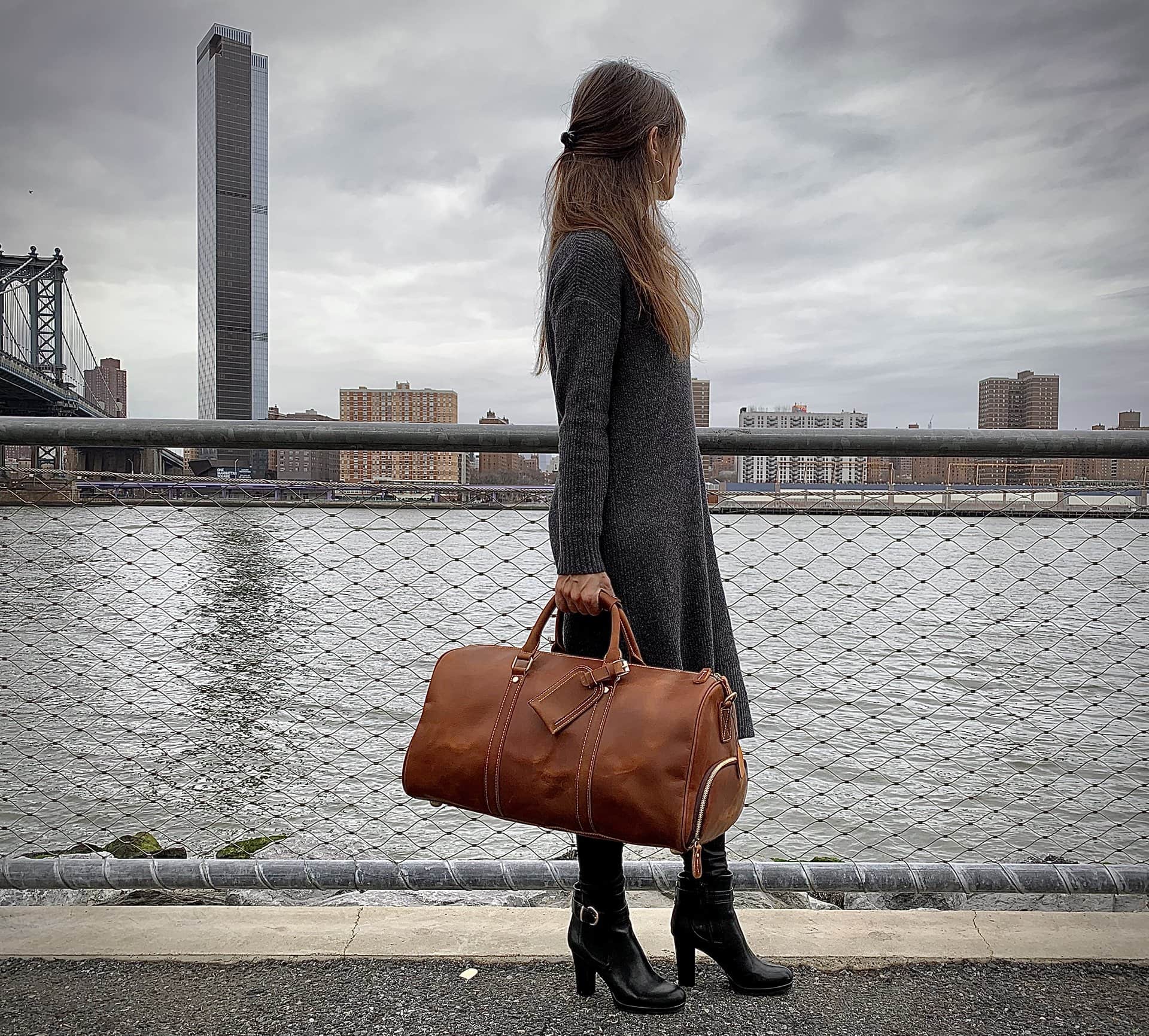 Woman holding a brown leather bag by a waterfront with a city skyline in the background