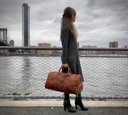 Woman holding a brown leather bag by a waterfront with a city skyline in the background