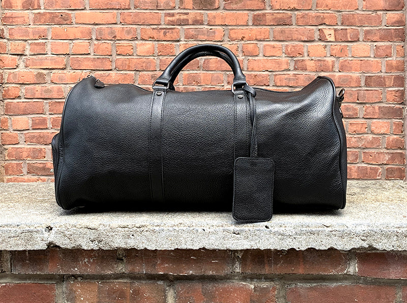 Black leather duffel bag with a tag on a stone ledge against a brick wall.