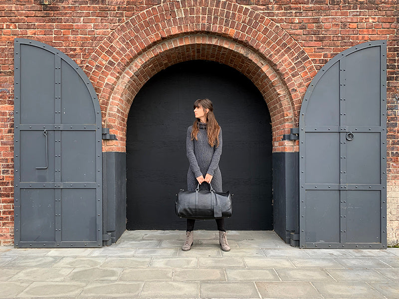 Person holding a black leather bag in front of a brick archway with large doors.