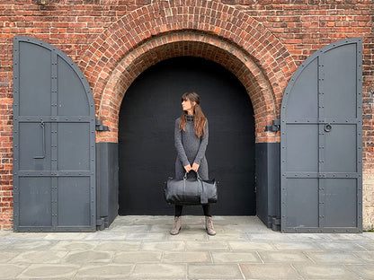 Person holding a black leather bag in front of a brick archway with large doors.