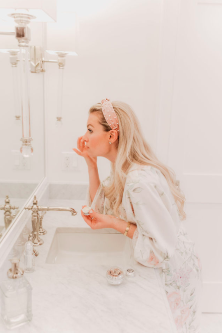 Woman applying makeup in a bathroom with a mirror and toiletries.