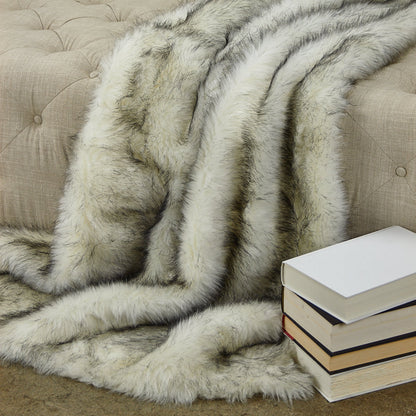 Gray and white fur throw blanket draped over a beige sofa with a stack of books on a wooden floor.