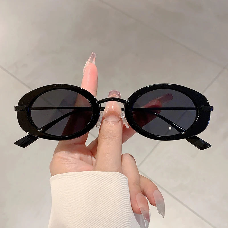 Black oval sunglasses held by a hand with clear nail polish against a neutral background