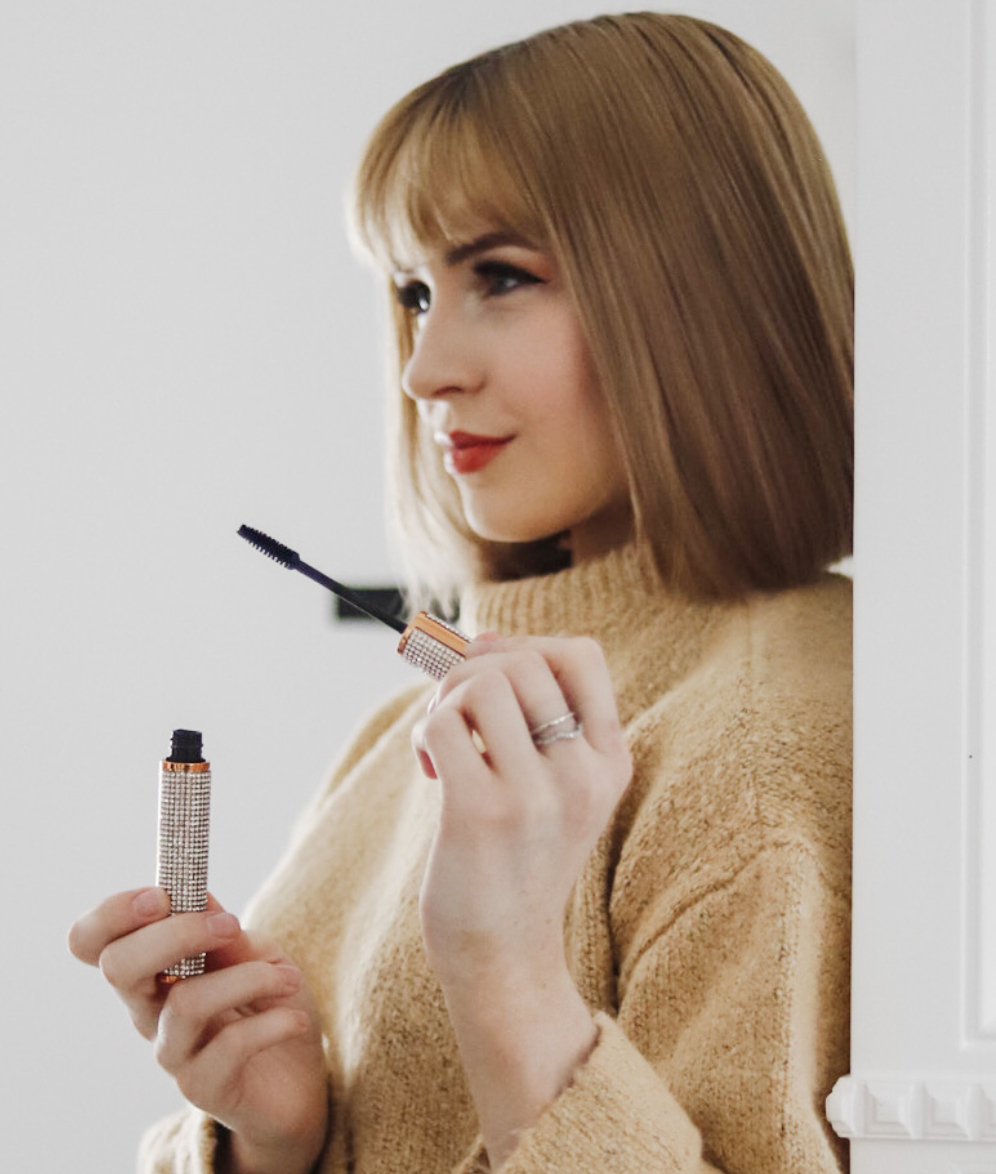 Woman holding a compact mirror and holding a Dr.  Liia bling mascara, wearing a beige sweater.