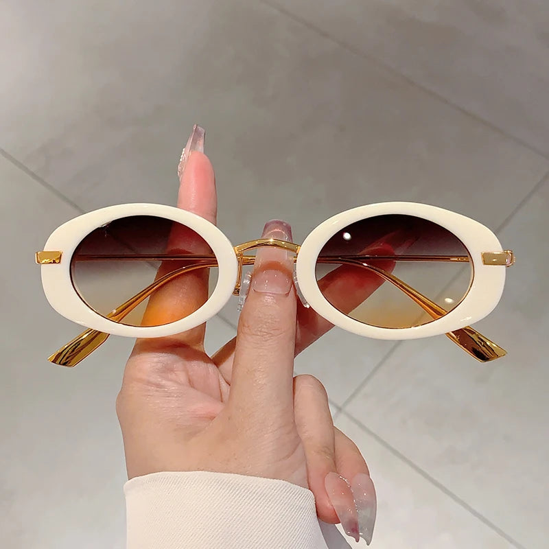 Hand holding a pair of sunglasses with gold frames and brown lenses against a neutral background
