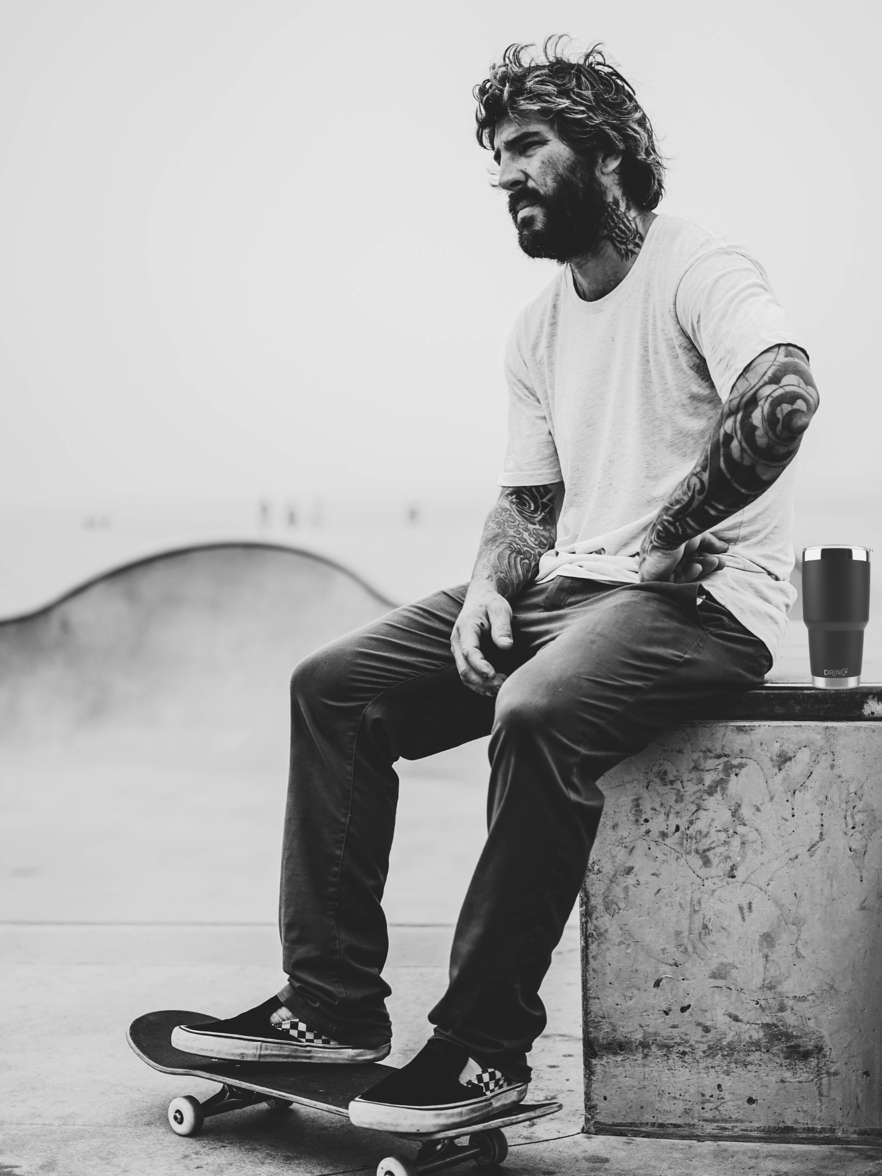 Man with tattoos sitting on a ledge with a skateboard, wearing a white t-shirt and dark pants with his DrinCo Tumbler.