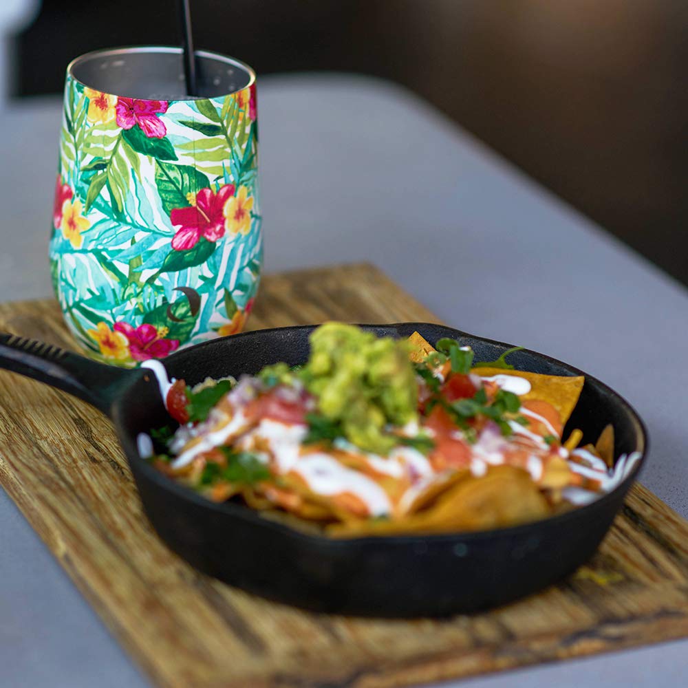 Skillet of nachos with guacamole on a wooden board next to a colorful floral-patterned cup.