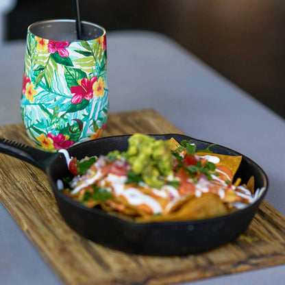 Skillet of nachos with guacamole on a wooden board next to a colorful floral-patterned cup.