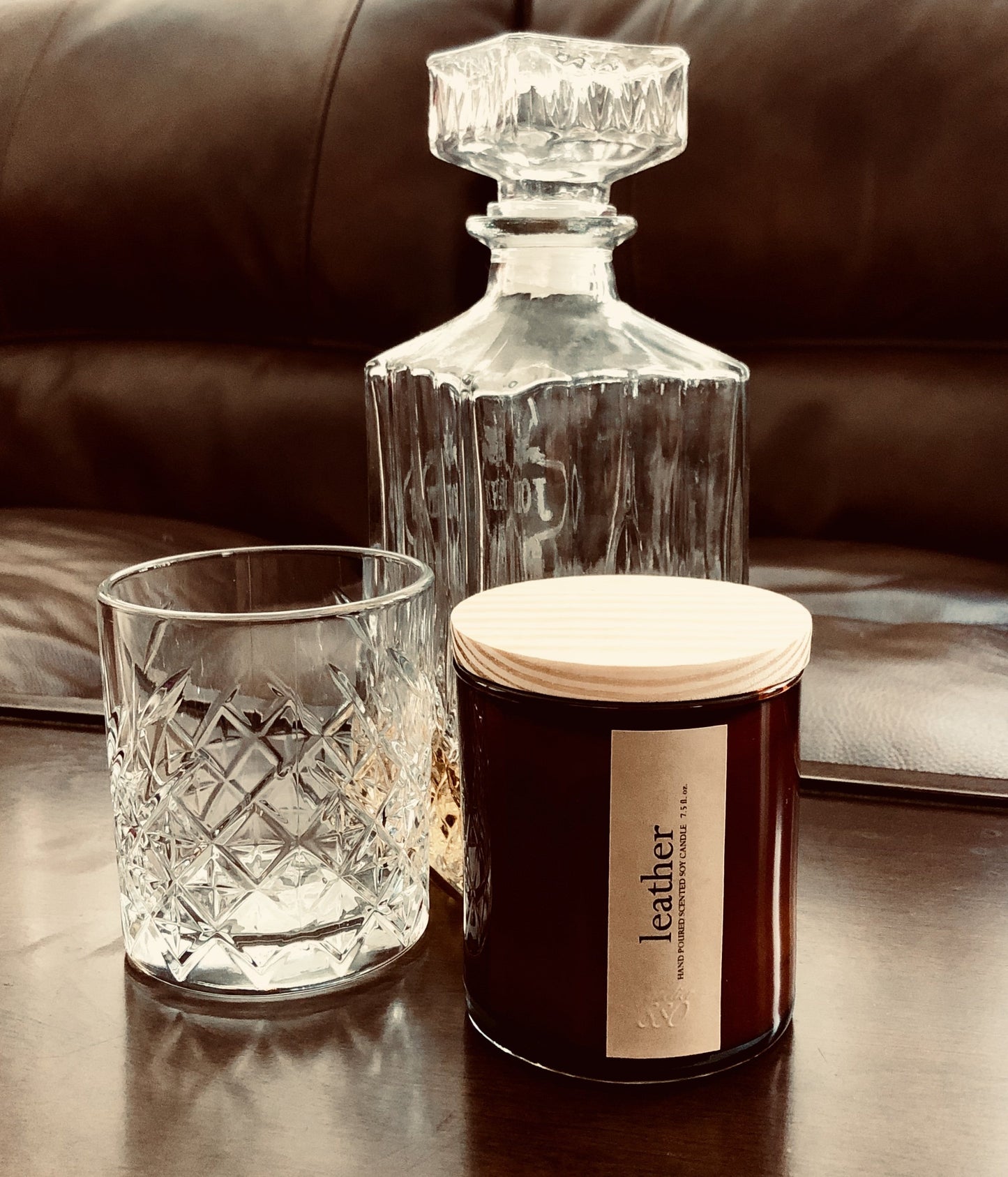 Glass decanter, tumbler, and candle labeled 'leather' on a reflective surface with a brown leather background.